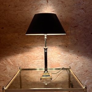 Könnte beinhalten: Ein schwarzer Lampenschirm sitzt auf einer Messing- und schwarzen Lampe. Die Lampe steht auf einem Beistelltisch mit Glasplatte und Messingverkleidung. Der Lampensockel ist quadratisch, und der Schirm hat eine klassische konische Form. Das Netzkabel ist sichtbar.