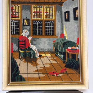 Könnte beinhalten: Gerahmtes Gobelin-Kunstwerk, das eine Innenszene mit einer lesenden Person darstellt. Das Kunstwerk zeigt eine Person in einer roten Jacke und weißer Kopfbedeckung, die in einem Raum mit einem Fenster und einem Bett sitzt. Die Farbpalette umfasst Braun, Rot, Grün und Gold.