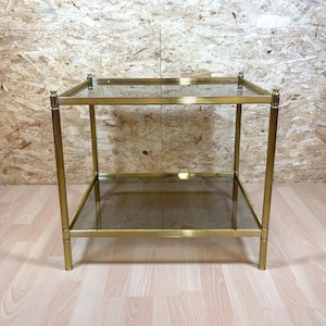 May include: A vintage brass side table with two glass shelves. The table has a rectangular shape and measures approximately 24 inches long, 18 inches wide, and 22 inches high. The brass frame has a polished finish, and the glass shelves are clear.