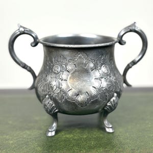 Könnte beinhalten: Eine silberfarbene, antike Zuckerdose mit zwei verzierten Griffen und drei kleinen Füßen. Die Schale weist detaillierte Blumenmuster und ein dekoratives Design auf.