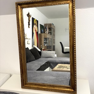 Könnte beinhalten: Ein rechteckiger Wandspiegel mit einem dicken, verzierten Goldrahmen und einem perlenartigen Innenrand. Die reflektierende Glasoberfläche zeigt einen klaren Blick in einen Raum mit einem grauen Sofa, einem Holztisch und einem Wandbehang.
