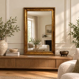 May include: A rectangular wall mirror with a thick, ornate gold frame featuring a beaded inner edge. The mirror reflects a bright living room interior with a white sofa, a fireplace, and a window. The frame has a distressed, metallic finish.