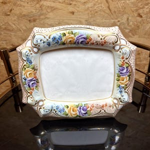 Könnte beinhalten: Ein verzierter, rechteckiger Bilderrahmen mit weißer Mitte und floralem Design. Der Rahmen zeigt gelbe, violette und blaue Rosen mit goldenen Akzenten auf cremefarbenem Hintergrund. Der Rahmen wird auf einer reflektierenden Oberfläche präsentiert.