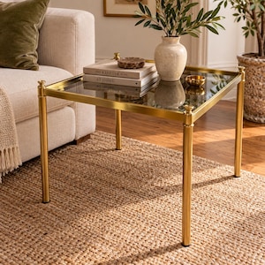 May include: A square side table with a polished gold metal frame and a clear glass top. The table holds two stacked books titled The Kinfolk Home and At Home, a small round brown decorative bowl, and a beige ceramic vase with green foliage.