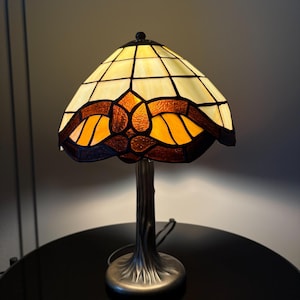 Könnte beinhalten: Eine Tischlampe aus Buntglas mit cremefarbenem und bernsteinfarbenem Schirm. Der Schirm hat ein geometrisches Muster mit einem Blumendesign in der Mitte. Der Sockel ist aus dunklem Metall mit einem Baumstammdesign.