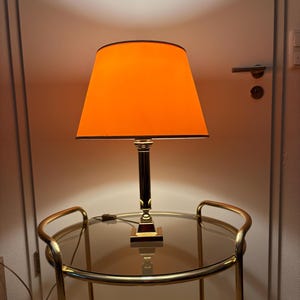 Könnte beinhalten: Eine Tischlampe mit orangefarbenem Lampenschirm und schwarzem Rand. Die Lampe hat einen goldfarbenen Sockel und Stiel. Sie steht auf einem Servierwagen mit Glasplatte und goldenem Rahmen. Die Lampe ist eingesteckt.