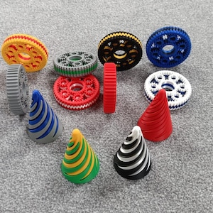 May include: An assortment of colorful 3D-printed gears and cone-shaped objects. The gears are in various colors, including yellow, green, red, black, blue, and white. The cones have striped patterns in multiple colors. The objects are arranged on a gray surface.