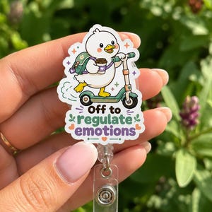 Op de afbeelding: Een witte eend badgehouder met een cartoon eend met een groene rugzak die een koffiebeker vasthoudt terwijl deze op een groene step rijdt. De tekst luidt Off to regulate emotions in zwarte en paarse letters, met kleine oranje en groene hartjes.