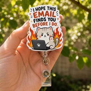 Può includere: Un portabadge con un adesivo sagomato raffigurante un gatto grigio davanti a un computer portatile circondato da fiamme arancioni. Il testo recita, I HOPE THIS EMAIL FINDS YOU BEFORE I DO. Il gatto ha un'espressione imbronciata e una zampa alzata.