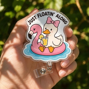 Pode incluir: Um porta-crachá de acrílico transparente com um pato de desenho animado com um laço rosa, sentado numa boia de flamingo rosa em água azul. O pato segura uma bebida amarela com uma fatia de limão. O texto JUST FLOATIN' ALONG está impresso em preto.