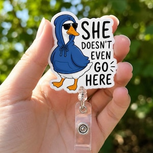 Pode incluir: Um porta-crachá com um pato branco vestindo um moletom azul com capuz e óculos de sol pretos. O texto diz SHE DOESN'T EVEN GO HERE em letras maiúsculas pretas. O pato tem um bico laranja e pés laranjas, fixado a uma tira de plástico.