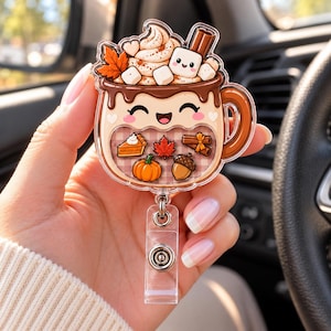 May include: An acrylic badge reel shaped like a mug of hot cocoa with a smiling face, topped with whipped cream, marshmallows, and a cinnamon stick. The mug features a shaker compartment containing miniature autumn icons like a pumpkin, leaf, and acorn.
