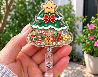 Op de afbeelding: Een kerstboomvormige badgehouder met een lachende gouden ster bovenop, een rode strik en een doorzichtige basis gevuld met feestelijke confetti. In het doorzichtige gedeelte zitten miniatuurbedels van een groene kerstbal, een zuurstok, een peperkoekmannetje, een cadeau en een ster.
