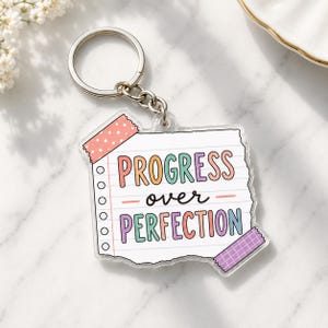 May include: A clear acrylic keychain shaped like a piece of lined notebook paper. The paper features the text PROGRESS over PERFECTION in colorful letters. A pink polka-dot piece of tape is at the top left and a purple grid tape is at the bottom right.