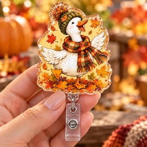 May include: An acrylic badge reel featuring a white duck wearing a green knit hat and a plaid scarf, standing amidst fallen autumn leaves. The duck has orange feet and an open beak. The design is set against a yellow background with maple leaves.