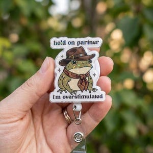 May include: A white badge reel with a cartoon frog wearing a brown hat and scarf. The text "hold on partner" is above the frog, and "I'm overstimulated" is below. The frog is green with glitter.