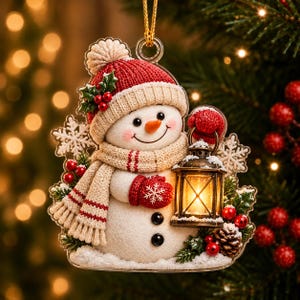 May include: A flat acrylic Christmas ornament featuring a smiling snowman wearing a red knit hat and a beige scarf. The snowman holds a lit lantern and is decorated with holly, pinecones, and white snowflakes on a clear background.