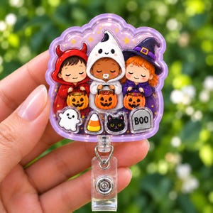 May include: A purple-rimmed badge reel featuring three children in Halloween costumes: a red devil, a white ghost, and a purple witch. Each holds a jack-o'-lantern bucket. Below them are a small ghost, a candy corn, a black cat, and a tombstone with BOO.
