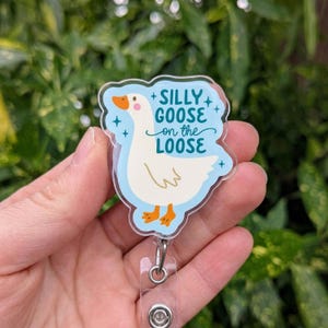 May include: A clear acrylic badge reel featuring a white goose illustration with orange beak and feet, set against a light blue background. The text "Silly Goose on the Loose" is written above the goose. The badge reel has a clear plastic strap and a metal clip.