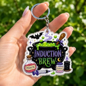 May include: A clear acrylic keychain featuring a black cauldron filled with bubbling green liquid, labeled "INDUCTION BREW" in purple and green text. It includes illustrations of a bat, a wooden spoon, a "PITOCIN" bottle, a syringe, a "CYTOTEC" pill bottle, and a black candle.