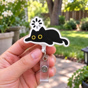 May include: A badge reel featuring a flat, black cat illustration with wide yellow eyes and a white loading icon above its head. The cat is attached to a clear plastic retractable cord mechanism with a metal clip and a clear vinyl strap attachment.