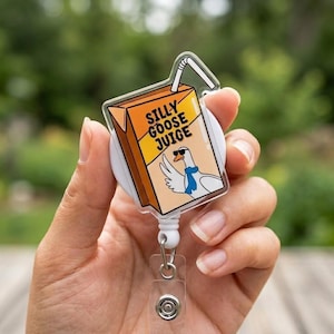 Puede incluir: Un portatarjetas retráctil blanco con una correa de plástico transparente y un diseño de caja de jugo de dibujos animados. La caja de jugo es naranja y marrón con el texto "SILLY GOOSE JUICE" y un ganso de dibujos animados con gafas de sol y una bufanda azul.