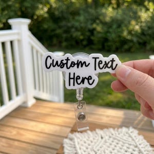May include: A white badge reel with the text "Custom Text Here" in black script. The badge reel has a clear plastic strap and a retractable cord. The badge is being held up in front of a blurred green background.