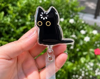 May include: A black cat badge reel featuring a glossy finish, two large yellow circular eyes, and a white loading icon graphic on the forehead. The cat-shaped acrylic piece is attached to a clear plastic strap with a metal swivel clip.