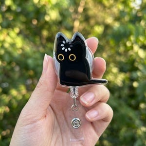 May include: A black cat-shaped badge reel with a clear plastic body. The cat has yellow eyes and a white floral design on its head. The reel has a retractable cord and a belt clip.