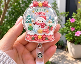 Op de afbeelding: Een ronde badgehouder van acryl met een sneeuwbolontwerp, voorzien van een sneeuwpop met een rode muts en sjaal, groene kerstbomen en cadeautjes. De tekst LET IT SNOW staat bovenaan. De basis is roze met een rode strik en gele ster.