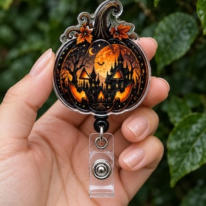 May include: A circular acrylic badge reel featuring a detailed illustration of a carved pumpkin with a spooky haunted house scene inside. The design includes a large orange moon, bare trees, and bats against a dark background with autumn leaves.