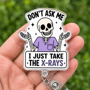 May include: A clear acrylic badge reel featuring a cartoon skeleton wearing a light purple scrub top with a pocket. The skeleton has its arms raised in a shrug. The text reads DON'T ASK ME I JUST TAKE THE X-RAYS in black and purple lettering.