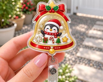 Op de afbeelding: Een doorzichtige acryl badgehouder in de vorm van een kerstklok, met een cartoonpinguïn die een kerstmuts en sjaal draagt. De klok is versierd met een rode strik, hulst en kleine feestelijke iconen zoals sneeuwvlokken, cadeaus en een boom.