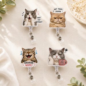 May include: Four retractable badge reels featuring cat illustrations with text. Top left: gray cat with 'WHAT JUST HAPPENED?!'. Top right: brown cat with 'NOT TODAY, HUMAN.'. Bottom left: tabby cat with 'HAHAHA NOPE!'. Bottom right: gray cat with 'I NEED COFFEE & SPACE'.