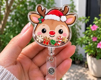 Op de afbeelding: Een doorzichtige acryl badgehouder met een cartoon rendierkop die een rode kerstmuts draagt. Het rendier heeft een rode neus en is omringd door kleine, losse feestelijke bedeltjes, waaronder een sneeuwvlok, een cadeautje en een zuurstok.