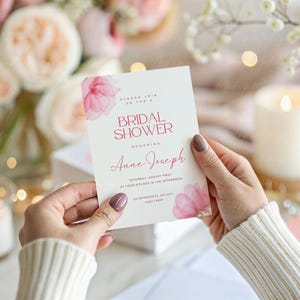 May include: A pink and white bridal shower invitation with watercolor flowers and the text "Please join us for a Bridal Shower Honoring Anne Joseph Saturday, August First at Four O'Clock in the Afternoon The Residence of Amy Smith 123 Example Way, Any City, Any State 12345"