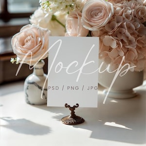 May include: A white card with the word "Mockup" in script is displayed in a bronze-colored holder. The card is in front of a bouquet of pink roses and white flowers. The text "PSD / PNG / JPG" is below the word "Mockup".