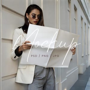 May include: A woman in a white blazer and striped pants is holding a blank white sheet of paper with the text "Mockup PSD / PNG JPG" printed on it.