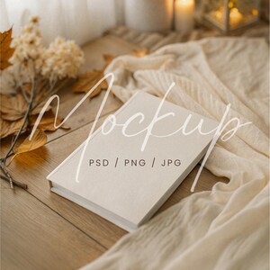 Puede incluir: Maqueta de libro con una cubierta color crema sobre una superficie de madera, drapeada con una tela color crema. Hojas y flores secas se suman a la estética otoñal. El texto "Mockup" y "PSD / PNG / JPG" son visibles.