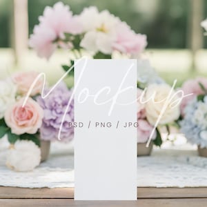 May include: A white rectangular card with the word "Mockup" in a script font, with "PSD / PNG / JPG" below it. The card is on a wooden table with floral arrangements in the background. The flowers are in shades of pink, purple, and white.