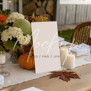 May include: A fall-themed table setting with a blank white sign. The table is decorated with a pumpkin, candles, and a floral arrangement with orange, white, and green flowers. A single brown leaf rests on the table.