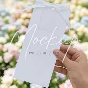 May include: A white rectangular card with a light blue ribbon tied in a bow. The card has the word "Mockup" written on it, along with "PSD / PNG / JPG". The card is held in front of a blurred background of colorful flowers.