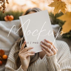 May include: A mockup book with a white cover is held in front of a person. The book is the focal point, with the word "Mockup" and "PSD / PNG / JPG" overlaid. The scene has an autumn theme, with fall leaves and a cozy sweater.