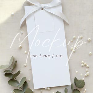 May include: A white rectangular card with a small tag, tied with a white ribbon. The card is surrounded by pearls and eucalyptus leaves. The word "Mockup" is written on the card, along with "PSD / PNG / JPG".