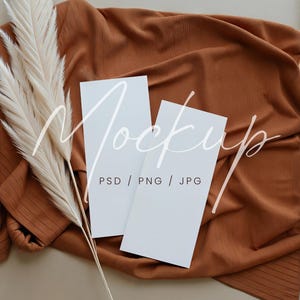 May include: Two blank white cards on a brown fabric background with a white pampas grass sprig. The text "Mockup PSD / PNG / JPG" is written on the cards.