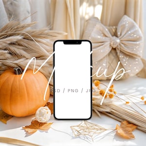 May include: A mockup image featuring a black smartphone with a blank white screen, surrounded by autumn-themed decorations. An orange pumpkin, dried wheat, and fall leaves are arranged with a decorative bow and a gold star-shaped ornament. The image includes the text "Mockup".