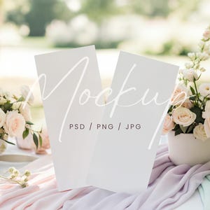 May include: Two white vertical cards with the word "Mockup" in a script font. The cards are on a table with pastel-colored fabric and floral arrangements. The text "PSD / PNG / JPG" is below the word "Mockup".