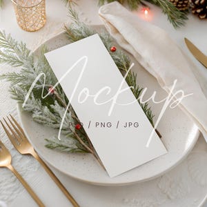 May include: A festive table setting with a white plate, gold cutlery, and a folded linen napkin. The plate holds a white card with text, adorned with frosted greenery and red berries. A gold candle holder and pine garland with lights complete the holiday theme.