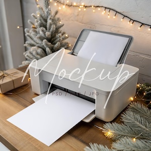 May include: A white printer with a sheet of paper emerging, placed on a wooden table. A small wrapped gift and a decorative Christmas tree are nearby. String lights and a brick wall are in the background.