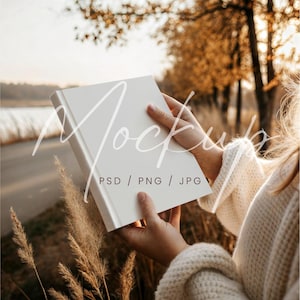 Könnte beinhalten: Ein leeres, weißes Buch im Freien gehalten, mit dem Text "PSD / PNG / JPG" sichtbar. Das Buch wird von einer Person gehalten, die einen cremefarbenen Pullover trägt. Der Hintergrund zeigt eine Straße, Bäume und hohes Gras mit warmem, goldenem Licht.